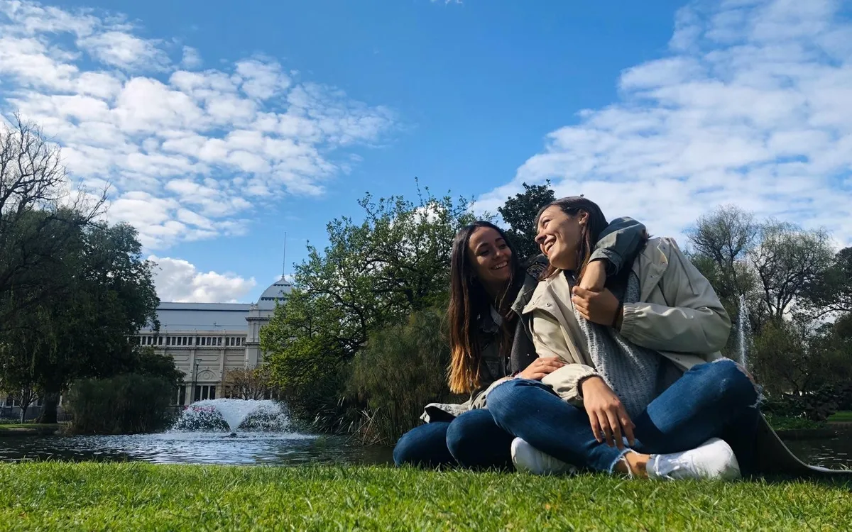 Dos estudiantes riendo sentadas en el césped de los Carlton Gardens, en Melbourne