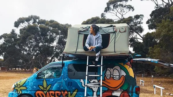 Estudiante sentada en la tienda de campaña del techo de una furgoneta de colores en Australia