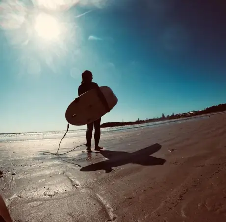 Estudiante con su tabla de surf caminando por la orilla, a contraluz