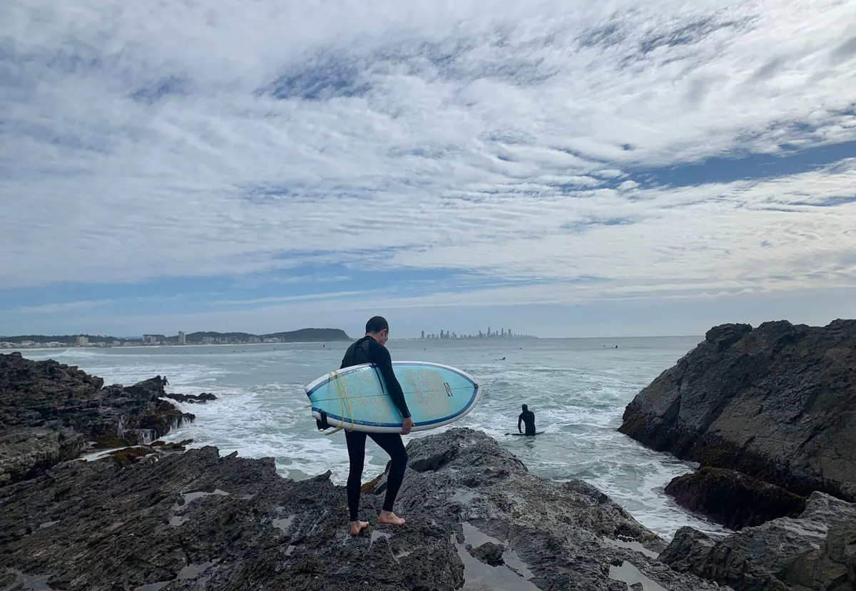 Un surfista con su tabla en las rocas de Snapper Rocks, con Gold Coast al fondo